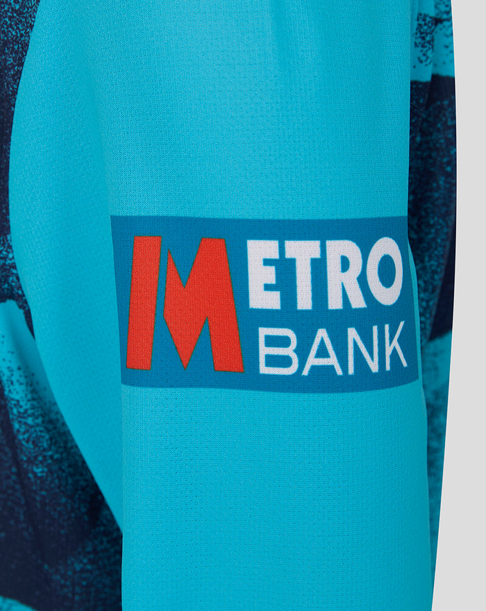 Close-up of turquoise sleeve with Metro Bank logo patch on the Junior 26/27 OD Long Sleeve Jersey.